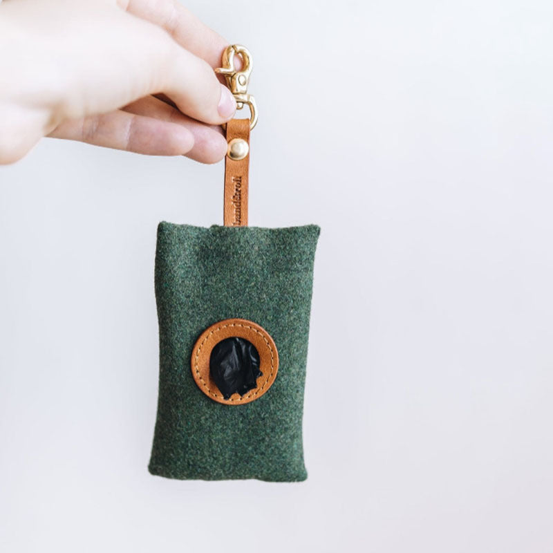 A portable green dog poop bag holder with a brown leather strap and a circular window revealing a black bag inside.