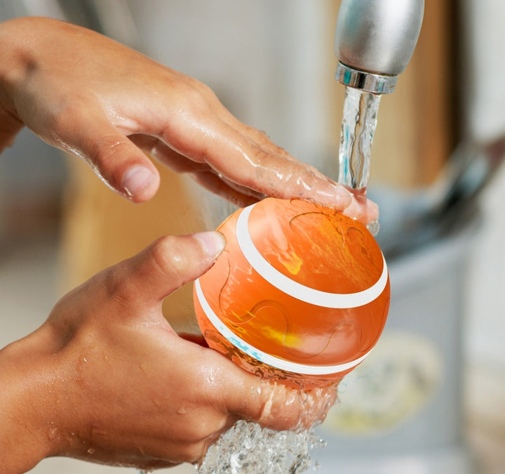Person washing an orange ball under running water from a faucet.