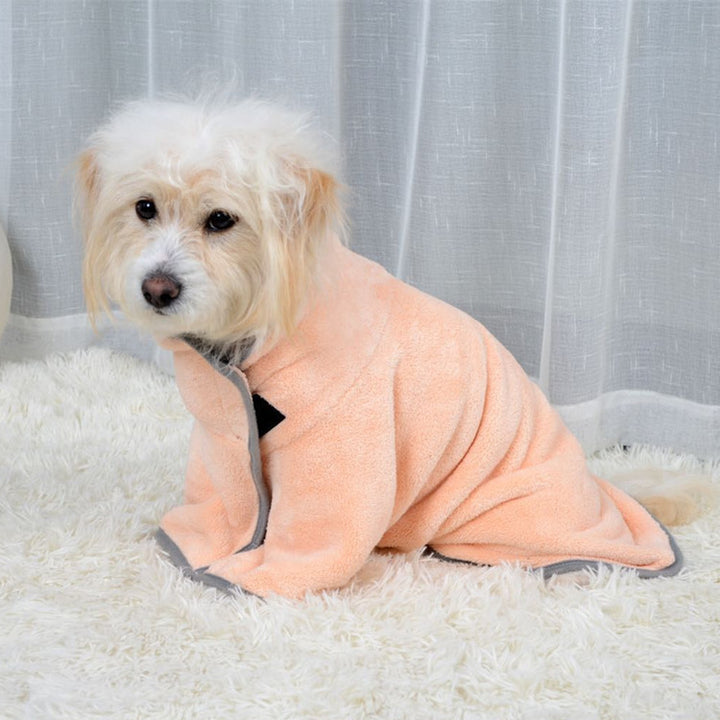 Dog wearing a peach-colored robe sitting on a white surface with a light gray curtain background